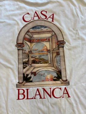 Vit t-shirt från Casablanca - Vit t-shirt från Casablanca med stort tryck på ryggen som föreställer en klassisk arkad med landskapsmotiv och röd text. T-shirten har en avslappnad passform och är tillverkad i mjuk bomull. Perfekt för dig som gillar unika prints och streetwear vibes.