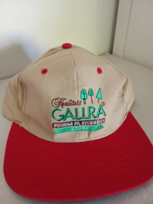 Ny keps  - Snygg keps i beige med röd skärm och broderad text 'Kvalitets Gallra' framtill. Kepsen har gröna och bruna detaljer samt ventilationshål i rött. Perfekt accessoar för en avslappnad stil.