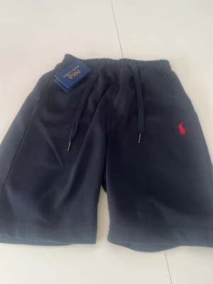 Marinblå shorts från Polo Ralph Lauren - Snygga marinblå shorts från Polo Ralph Lauren med elastisk midja och snörning. Klassisk röd logga broderad på vänster ben. Tillverkade i mjuk bomull för maximal komfort. Perfekta för sommardagar och avslappnade tillfällen.