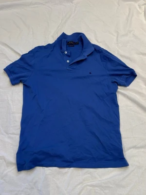Blå pikétröja från Ralph Lauren - Klassisk blå pikétröja från Ralph Lauren med korta ärmar och krage. Tröjan har två knappar vid halsen och en liten broderad logga på bröstet. Perfekt till jeans eller shorts för en avslappnad stil.