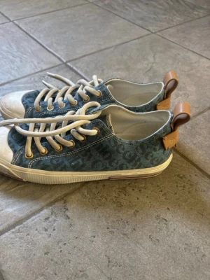 Blå By Chloé sneakers med monogram - Snygga blå sneakers från By Chloé med monogrammönster i tyget, vita snören och tå, samt bruna detaljer i läder baktill. Skorna har en klassisk låg profil och vit sula som ger en fräsch look till din outfit.