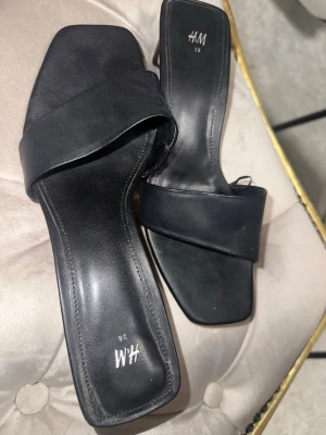 Svarta slip-in sandaler från H&M - Stilrena svarta slip-in sandaler från H&M med bred rem och fyrkantig tå. Tillverkade i konstläder med minimalistisk design som passar perfekt till jeans eller klänning. Snygg och enkel modell för dig som gillar trendiga detaljer. 