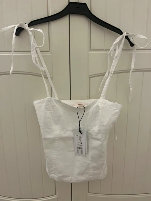 Vit linnetopp med knytband - Snygg vit linnetopp med smala axelband som knyts på axlarna. Toppen har en rak, croppad passform och är tillverkad i bomull. Perfekt för varma dagar och enkel att styla med jeans eller kjol.