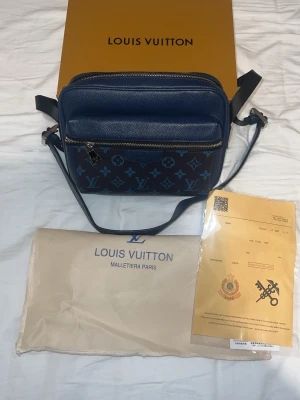 Blå axelväska från Louis Vuitton - Snygg blå axelväska från Louis Vuitton med klassiskt monogrammönster i olika blå nyanser. Väskan har justerbar axelrem, silverfärgade dragkedjor och är tillverkad i tåligt läder. Perfekt för dig som vill ha en exklusiv och trendig accessoar.