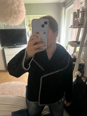 Svart kappa med kontrastsömmar - Snygg svart kappa med oversized krage och vita kontrastsömmar längs kanterna. Kappan har en rak passform och stängs framtill. Materialet ser ut att vara mjukt tyg, perfekt för hösten/våren! I storlek 158/164 men passar mig som har XS 