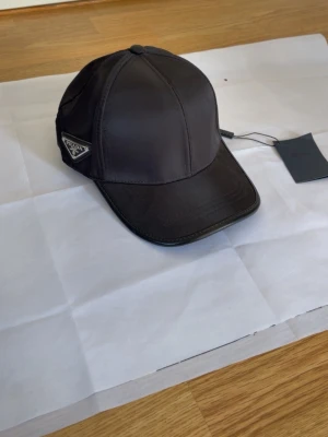 Svart keps från Prada i bomull - Snygg svart keps från Prada med klassisk böjd skärm och ikonisk metallogo på sidan. Tillverkad i 100% bomull för en bekväm känsla och justerbar passform. Perfekt accessoar för en trendig och stilren look.