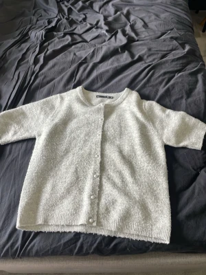 Vit kofta med pärlknappar - Mysig vit kofta med kort ärm och rund halsringning. Koftan har dekorativa pärlknappar framtill och är stickad i ett fluffigt material som ger en soft känsla. Perfekt att slänga över en topp för en chill look.