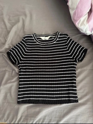 Svartvit ribbad randig topp - Snygg svart topp med vita horisontella ränder och ribbad struktur. Kortärmad och croppad modell med rund halsringning. Perfekt till jeans eller kjol för en trendig look.