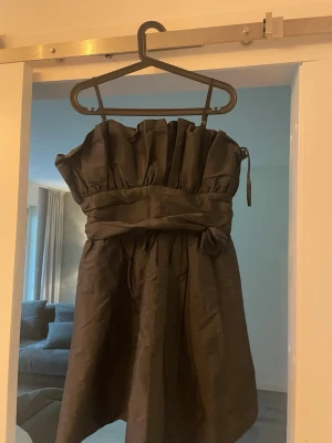 Svart axelbandslös klänning SWE-MIX - Snygg svart axelbandslös klänning från SWE-MIX med markerad midja och knytband. Klänningen har en kort längd och ett elegant, puffigt liv. Perfekt för dig som vill ha en stilren och chic look.