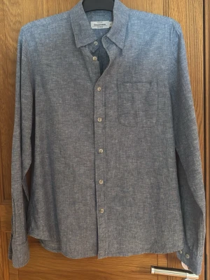 Blå linneskjorta från Dressmann - Stilren blå skjorta från Dressmann i mjukt linne. Skjortan har klassisk krage, bröstficka och knappar framtill. Långärmad modell med avslappnad passform, perfekt för en enkel och snygg look.