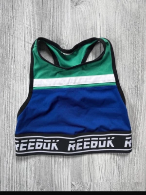 Reebok sport-bh blå och grön XXS (150) - Cool sport-bh från Reebok med blå och grön färgblock, vit rand och svart kant. Elastiskt band med Reebok-logga runt om. Racerback-design och bekvämt material som passar perfekt för träning och sport. Storlek barn 150 (XXs)