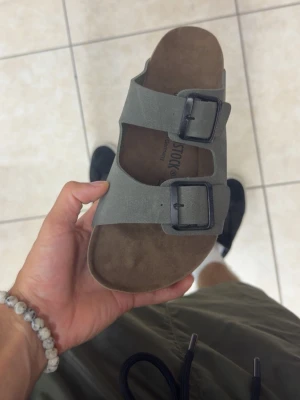 Birkenstock sandaler - Säljer ett par klassiska Birkenstock sandaler i grå mocka med två breda remmar och stora metallspännen. Fotbädden är i brunt läder och sulan är svart. Perfekta för dig som gillar stilren och bekväm design.