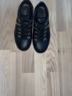 Svarta Balmain sneakers med metalldetaljer - Snygga svarta sneakers från Balmain med coola metalldetaljer och svarta skosnören. Skorna har en låg profil och är tillverkade i läder med Balmain-logga på plösen och insidan. Perfekta för dig som vill ha en stilren men edgy look.
