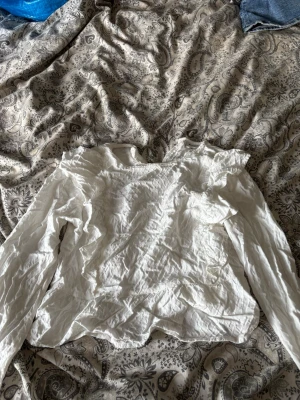 Vit volangblus med lång ärm - Säljer en vit blus med volangdetaljer och lång ärm. Blusen har en lätt skrynklig struktur och är tillverkad i ett tunt, luftigt tyg. Perfekt för dig som gillar romantiska och feminina plagg med extra detaljer vid axlar och ärmslut.