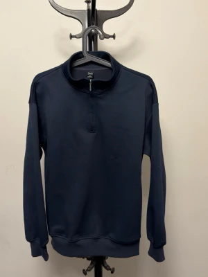 Mörkblå half-zip tröja från Dazy - Mörkblå långärmad tröja från Dazy med half-zip dragkedja vid halsen och ståkrage. Tröjan har ribbade muddar vid ärmslut och nederkant, och är tillverkad i ett mjukt material som känns bekvämt mot huden. Enkel och stilren design som passar till många outfits.