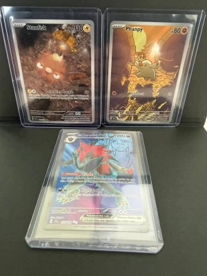 Pokémonkort Stunfisk, Phanpy & Zoroark - Tre Pokémonkort: Stunfisk med mörkbrun och gul design, Phanpy med orange och gul bakgrund samt Hisuian Zoroark VSTAR med färgstark illustration. Alla kort är i plastfickor och har detaljerade illustrationer och holografiska effekter.