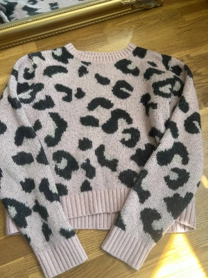 Rosa leopardmönstrad stickad tröja - Säljer en rosa stickad tröja med svart leopardmönster. Tröjan har rund halsringning, långa ärmar och ribbade muddar vid ärmslut och nederkant. Perfekt för dig som gillar djurmönster och vill ha en trendig look.