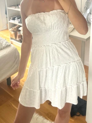 Vit axelbandslös sommarklänning - Söt vit axelbandslös klänning med broderade spetsdetaljer och smockad midja. Klänningen har volanger och är kort i modellen, perfekt för varma dagar. Tillverkad i bomull för en luftig och bekväm känsla.