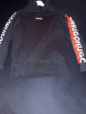 Mörkblå hoodie från Hugo med tryck - Mörkblå hoodie från Hugo med vit logga på bröstet och stora HUGO-tryck längs båda ärmarna, ena ärmen har röd panel. Klassisk huva och ribbade muddar. Perfekt för en streetwear-look och tillverkad i mjuk bomullsblandning passar xs .
