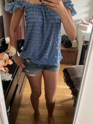 Blå volang topp med offshoulder - Säljer en blå topp med volangdetaljer och offshoulder-design. Toppen har en luftig och loose passning med rynkad kant upptill och volanger över hela plagget. Perfekt för dig som gillar en feminin och trendig stil.