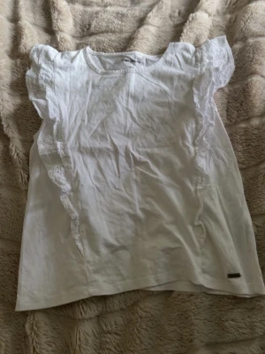Vit blus med volangdetaljer - Söt vit blus med volang längs axlar och sidor. Blusen är ärmlös och har rund halsringning. Tillverkad i ett mjukt bomullstyg som känns skönt mot huden. Perfekt för dig som gillar en feminin och enkel stil.