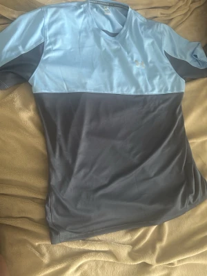Blå och grå t-shirt från Under Armour - Snygg tvåfärgad t-shirt från Under Armour i ljusblått och mörkgrått. Tillverkad i ett lätt och funktionellt syntetmaterial som andas, perfekt för träning. Klassisk passform med korta ärmar och rund halsringning. Diskret logga på bröstet.