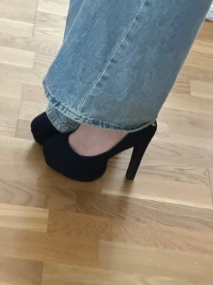 Svarta klassiska pumps med platå 39 - Säljer ett par svarta pumps med hög klack och platå framtill. Skorna har rundad tå och är tillverkade i ett tygmaterial som ger en matt finish. Perfekta för dig som vill ha en tidlös och stilren look.