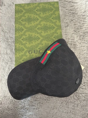 Svart Gucci keps med GG-mönster - Snygg svart keps från Gucci med klassiskt GG-monogram över hela kepsen. Har en grön och röd rand med guldbroderad bi på sidan. Justerbar rem bak i läder och insida i brunt tyg. Perfekt accessoar för dig som gillar lyxiga detaljer.