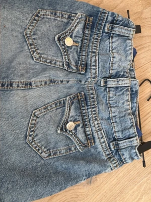 Snygg Blå jeanskjol till sommaren  - Snygg blå jeanskjol i kort modell med slitna detaljer och rå kant. Kjolen har klassiska fem fickor, bälteshällor och dekorativa sömmar framtill. Två bakfickor med lock och knapp för extra stil. Perfekt till en avslappnad look.
