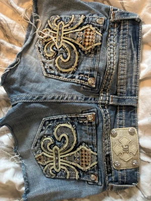 Miss Me jeansshorts med broderi - Säljer ett par blå jeansshorts från Miss Me med stora broderade bakfickor, glittriga detaljer och dekorativa nitar. Shortsen har låg midja och slitna kanter för en cool look i storlek XS - S. Perfekta för dig som gillar statement-plagg! Jag säljer pågrund av att de tyvärr blivit försmå. De är köpta på Plick som jeans men jag gjorde om de till shorts, väldigt fina och i väldigt bra skick. (Mina favoriter) 💗💗