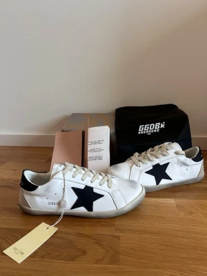 Vita Golden Goose sneakers med svart stjärna - Snygga vita sneakers från Golden Goose med svart stjärna på sidan och svart detalj på hälen. Skorna har vita snören och en klassisk låg siluett. Tillverkade i läder med slitstark gummisula. Perfekta för dig som gillar streetstyle och vill ha något trendigt.