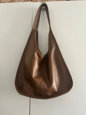 Brun axelremsväska i skinnimitation - Snygg brun handväska i skinnimitation med mjuk, slouchy form och bred axelrem. Väskan har dragkedja upptill och en enkel, minimalistisk design som passar till många olika stilar. Perfekt för dig som gillar trendiga accessoarer. Väskan är köpt secondhand och är lite slitig på insidan av bandet. 