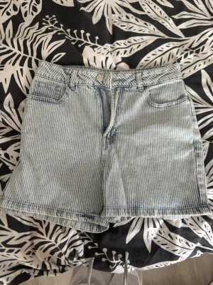 Randiga denimshorts med hög midja - Säljer ett par ljusblå denimshorts med vita smala ränder och hög midja. Klassisk femficksmodell med knapp och dragkedja framtill. Perfekta till sommaren och ger en avslappnad vibe.