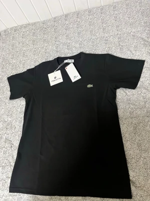 Svart t-shirt  - Snygg svart t-shirt med den klassiska gröna krokodilloggan broderad på bröstet. T-shirten har rund halsringning och korta ärmar. Tillverkad i mjuk bomull för en skön känsla och stilren look.
