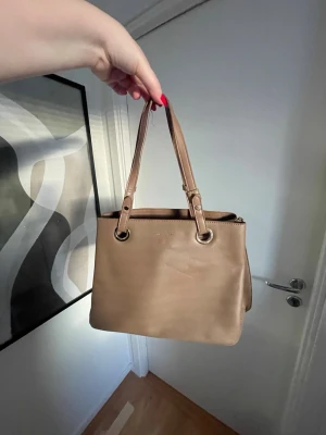 Beige handväska  - Stilren beige handväska från Jenny Fairy med silverfärgade metalldetaljer och en fyrkantig form. Perfekt för dig som gillar minimalistisk design och vill ha plats för det viktigaste. Finns en liten skråma framme på väskan, se första och sista bild 