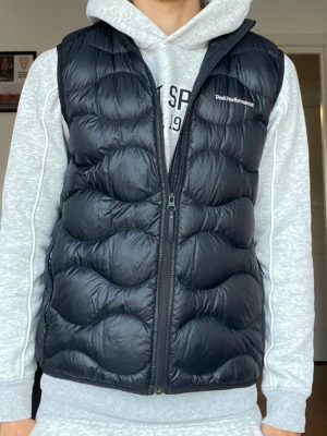 Svart dunväst Peak Performance - Snygg svart dunväst från Peak Performance med vågigt quiltat mönster och dragkedja framtill. Västen har hög krage och två sidofickor med dragkedja. Perfekt att slänga över en hoodie för extra värme. Klassisk logga på bröstet.