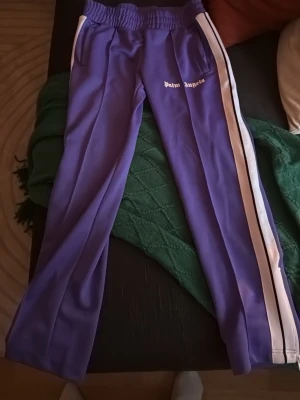 Lila mjukisbyxor från Palm Angels - Snygga lila mjukisbyxor från Palm Angels med vita sidoränder och logga på ena benet. Byxorna har resår i midjan och raka ben. Perfekta för en chill och trendig look.