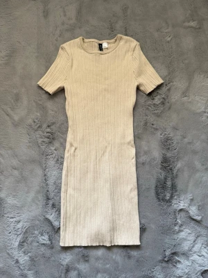 Beige ribbad kortklänning Divided - Säljer en beige ribbad kortklänning från Divided. Klänningen har korta ärmar, rund halsringning och är i ett mjukt, stretchigt material som sitter snyggt på kroppen. Perfekt för dig som gillar enkel och stilren design.