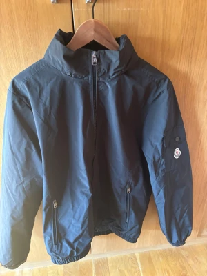 Svart vindjacka från Moncler - Snygg svart vindjacka från Moncler med hög krage och dragkedja framtill. Jackan har två fickor med dragkedja och en Moncler-logga på ärmen. Tillverkad i lätt och slitstarkt material, perfekt för blåsiga dagar. Så jävla stilig bra pris pågrund av att den är köpt på plick, man kan inte ens se att den är använd superpris 899