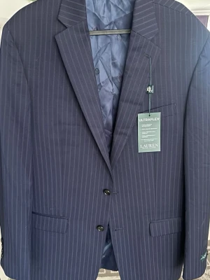 Mörkblå randig kavaj Lauren Ralph Lauren - Snygg mörkblå kavaj från Lauren Ralph Lauren med tunna vita kritstrecksränder. Klassisk modell med två knappar framtill, tre fickor och fyra knappar vid ärmslutet. Märkt med Ultraflex för extra komfort. Perfekt för en stilren look.