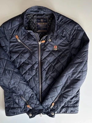 Morris Jacka - Snygg marinblå quiltad jacka från Morris med dragkedjor och läderdetaljer. Jackan har ett emblem på bröstet, ståkrage och innerfoder med prickigt mönster. Nypris : ca 2000 SEK Pris : 400 SEK
