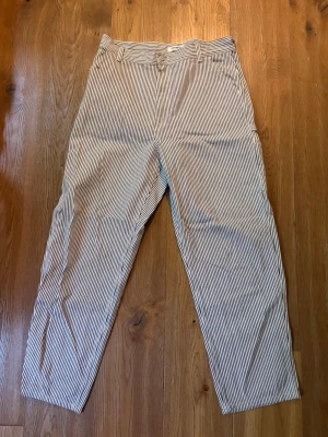 Carpenter pants från Carhartt - Snygga byxor med smala vertikala ränder i vitt och mörkblått. Byxorna har raka ben, normal midja och klassisk knappgylf. Perfekta för en avslappnad och stilren look. OBS! Fläck vid väster ficka, därav billigare pris 