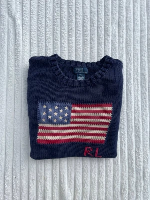 Mörkblå stickad tröja Polo Ralph Lauren - Mörkblå stickad tröja från Polo Ralph Lauren med amerikansk flagga och RL-broderi på bröstet. Tröjan har rund halsringning och ribbstickade muddar vid ärmslut och nederkant. Perfekt för dig som gillar klassisk preppy stil.
