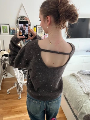 Mörkgrå stickad tröja med öppen rygg - Säljer en brun stickad tröja med glittriga detaljer och unik öppen rygg med band. Tröjan har avslappnad passform, långa ärmar och ribbade muddar. Perfekt för dig som vill ha något extra till vardagslooken.