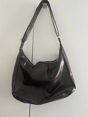 Mörkbrun axelväska i skinn - Snygg mörkbrun axelväska i glansigt skinn med bred axelrem och dragkedja upptill. Väskan har en mjuk, slouchy form och minimalistisk design utan synligt märke. Perfekt för dig som gillar stilrena accessoarer. Väskan är köpt secondhand. 