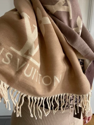 Beige-brun halsduk från  Vuitton - Snygg och lyxig halsduk från Vuitton i beige och brun med stora logotyper och text. Halsduken har dekorativa fransar i ändarna och är tillverkad i ett mjukt, vävt material som känns skönt mot huden. Perfekt accessoar för att lyfta din outfit cashmire