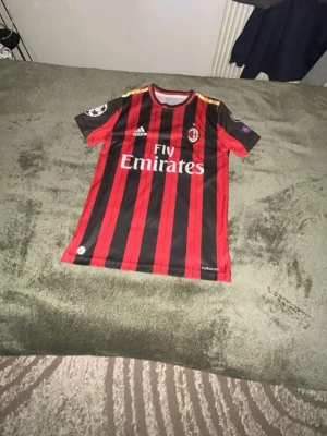 AC Milan matchtröja Kaka #22 - Snygg AC Milan matchtröja från Adidas med Kaka och nummer 22 på ryggen. Tröjan är röd och svart med vertikala ränder, Champions League-märke på ärmen och Fly Emirates-tryck på bröstet. Tillverkad i lätt och ventilerande material, perfekt för fotboll eller streetwear. Jag är 160cm lång och den passar perfekt på mig.