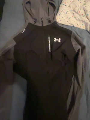 Svart och grå hoodie från Under Armour - Snygg svart och grå hoodie från Under Armour med dragkedja framtill och logga på bröstet. Hoodien har en justerbar huva och är tillverkad i ett mjukt, sportigt material som passar perfekt för träning eller chill. Raka ärmar och modern passform.