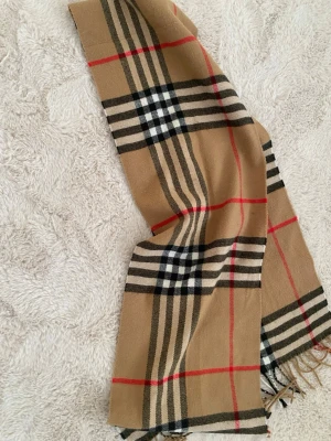 Beige halsduk med rutigt mönster - Snygg beige halsduk med klassiskt rutigt mönster i svart, vitt och rött. Halsduken har fransar i ändarna och är tillverkad i ett mjukt tyg som känns skönt mot huden. Perfekt accessoar för att lyfta din outfit.