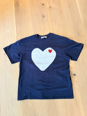 Blå Comme des Garçons Play t-shirt - Säljer en mörkblå t-shirt från Comme des Garçons Play med ett stort vitt hjärta och en liten röd hjärtapplikation på bröstet. T-shirten har klassisk passform och är kortärmad, tillverkad i mjuk bomull. Perfekt för dig som gillar coola designer och statement-plagg.